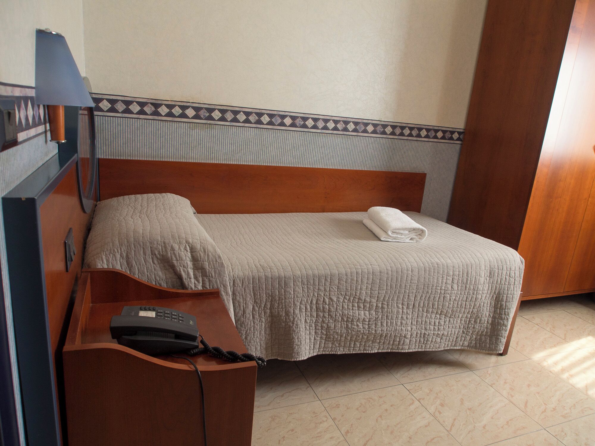 A hotel room with a single bed, a wooden headboard, a bedside table with a telephone, and a patterned wall.