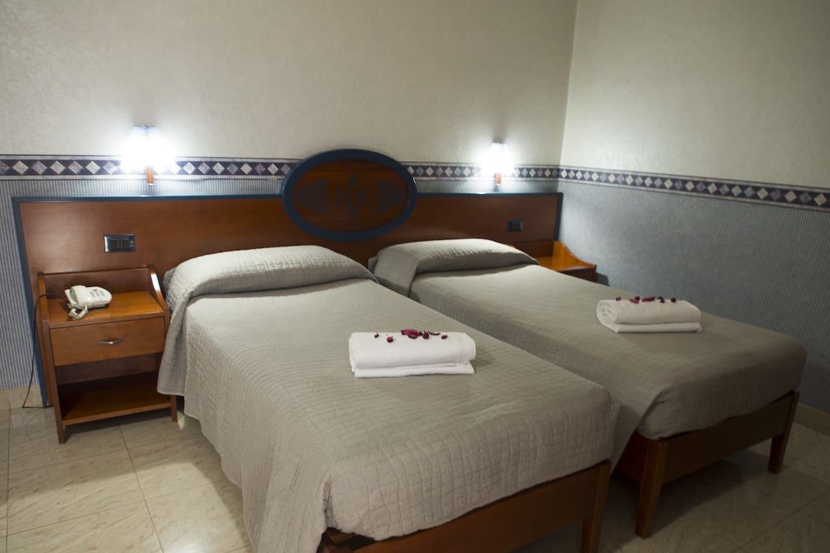 A hotel room with two beds, a nightstand with a phone, and a wall-mounted light fixture.