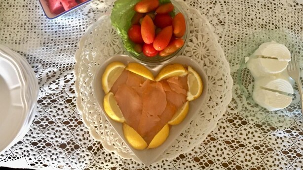 Une table recouverte d’une nappe en dentelle blanche, un saladier rempli de morceaux de pastèque, un autre saladier contenant des rondelles de carottes, une assiette avec du saumon et des tranches de citron, et un récipient en verre contenant une substance blanche.