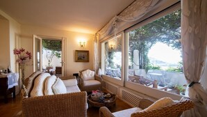 Un salon chaleureux avec des meubles en osier, un coin repas avec une table dressée pour deux personnes, et une grande fenêtre offrant une vue sur l’extérieur.