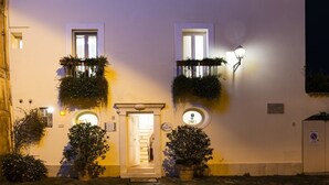 Un bâtiment à la façade blanche, doté de deux fenêtres ornées de plantes suspendues, d’une porte surmontée d’une lampe et d’une petite fenêtre circulaire également éclairée.