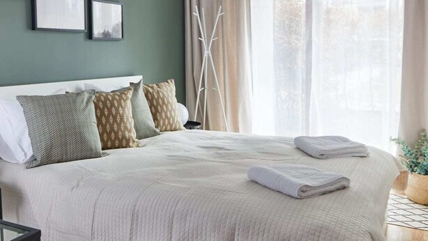 Een net opgemaakt bed met een witte quilt, een nachtkastje met een lamp, en een raam met dunne gordijnen.