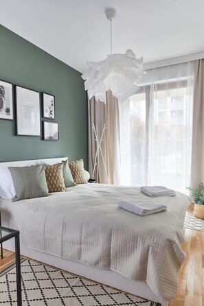 Un lit bien fait, recouvert d’une couette blanche, une table de chevet avec une lampe et une fenêtre ornée de rideaux légers.
