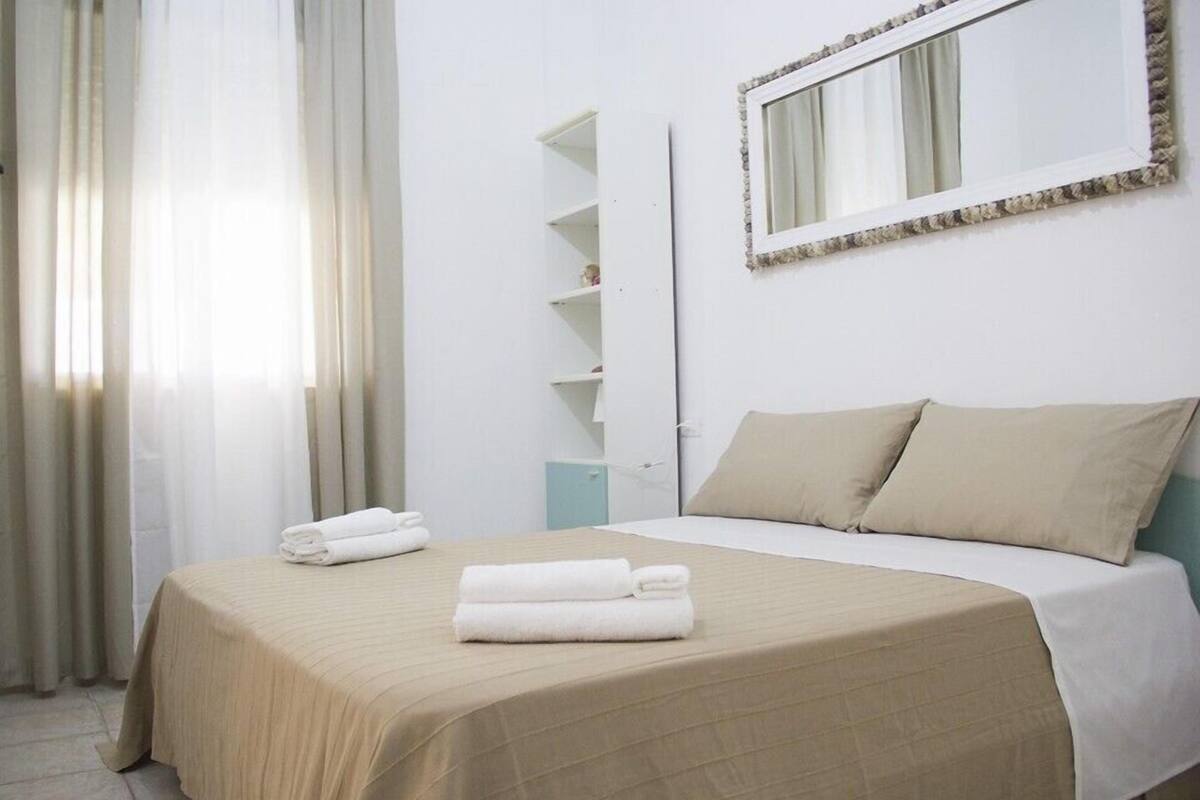 A neatly made bed with beige bedding and two pillows, a bedside table with a lamp, a mirror on the wall, and a window with sheer curtains.
