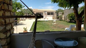 A view from a balcony with a stone wall, a small table, and chairs. In the background, there is a house with a tiled roof, a lawn, and a statue. - Cozzo Spadaro B&B (Pachino)