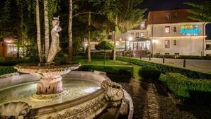 En veloplyst springvand i en anlagt have med en bygning med skiltet "Hotel Afrodyta" i baggrunden.
