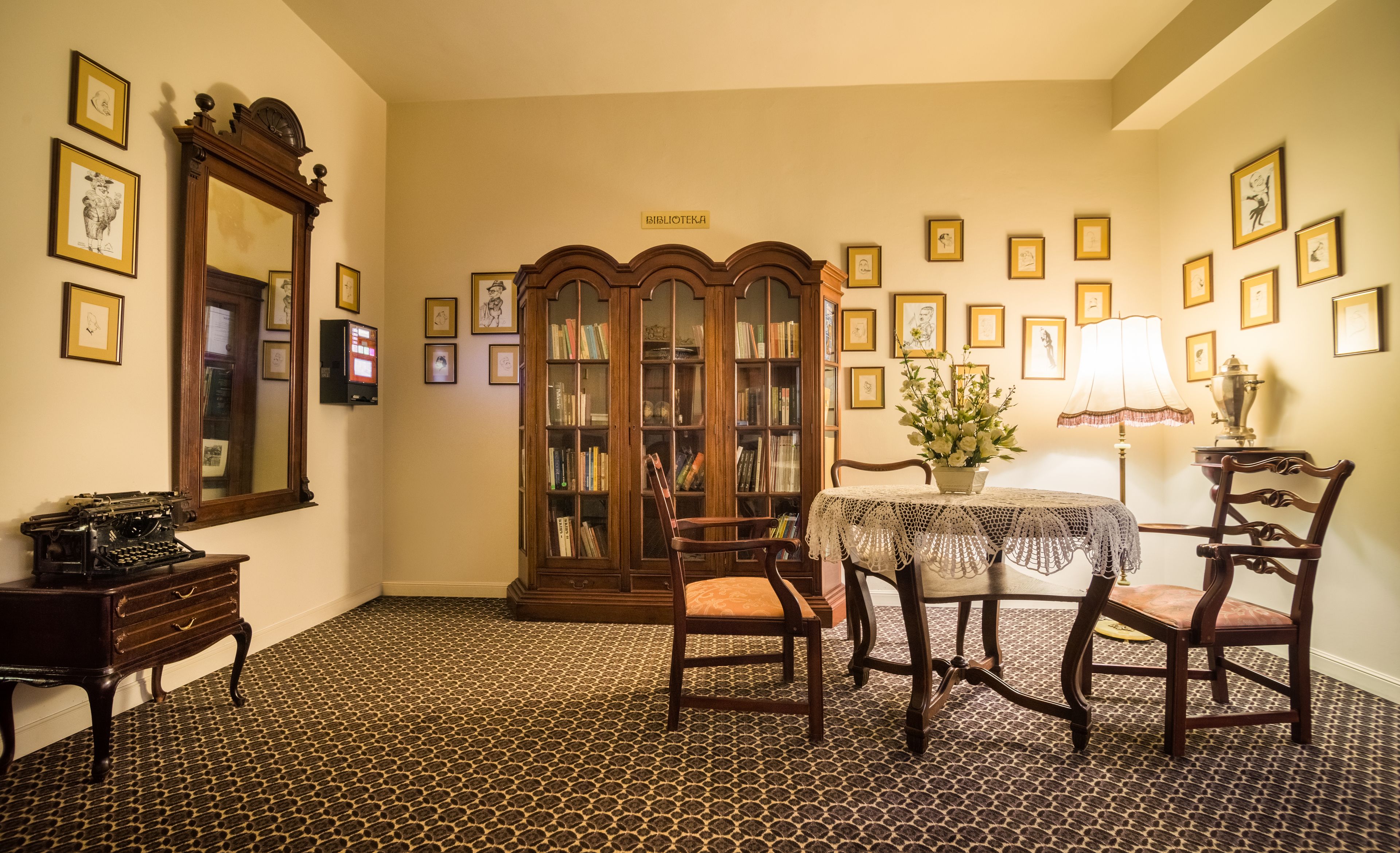 Um quarto com piso estampado, móveis de madeira, uma estante de livros e quadros nas paredes, com uma placa indicando "BIBLIOTEKA".