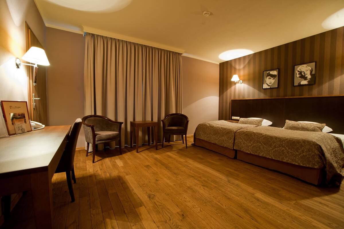 Habitación de hotel con piso de madera, un escritorio con silla, dos camas individuales, una mesita pequeña y dos lámparas de pared.