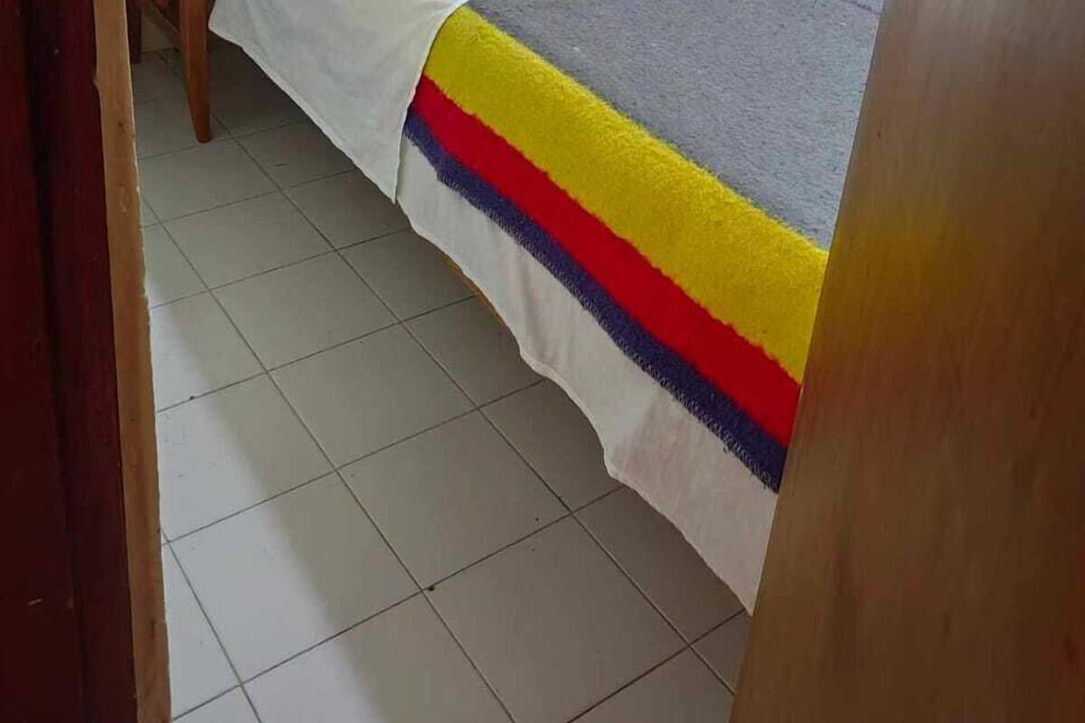 A small, single-bed room with a wooden door, a small table, and a bed with a colorful blanket.