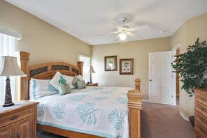 A bedroom with a bed, wooden headboard, ceiling fan, bedside lamps, framed pictures on the wall, and a door.