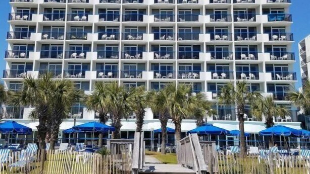 A multi-story hotel with balconies, a pool area with umbrellas, and a wooden staircase leading to the entrance. - Boardwalk 932 (Myrtle Beach)