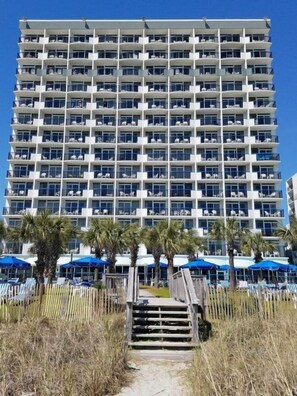 A multi-story hotel with balconies, a pool area with umbrellas, and a wooden staircase leading to the entrance. - Boardwalk 932 (Myrtle Beach)