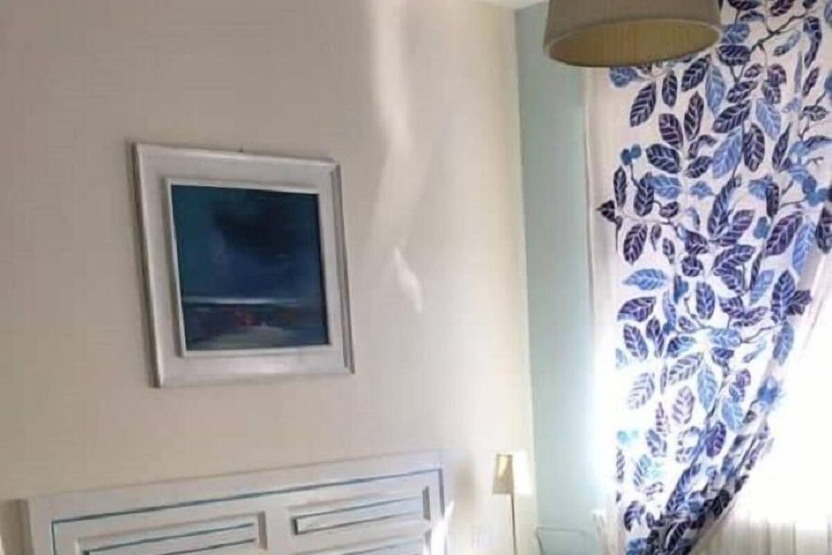 A bedroom with a bed, a bedside table, a lamp, a painting on the wall, and a window with curtains.