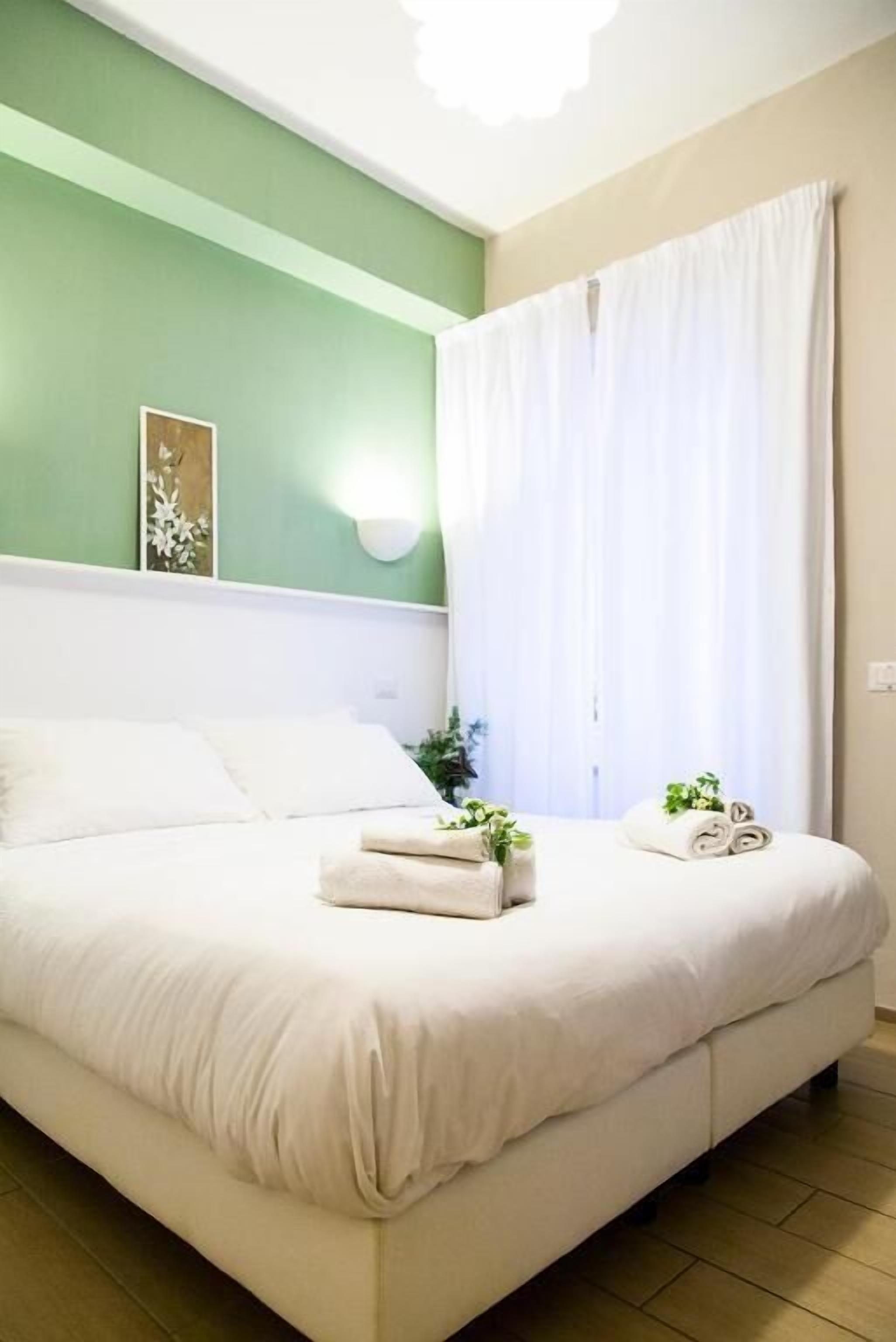 Una cama bien tendida con sábanas blancas y dos toallas enrolladas, un aplique en la pared y una ventana con cortinas blancas.