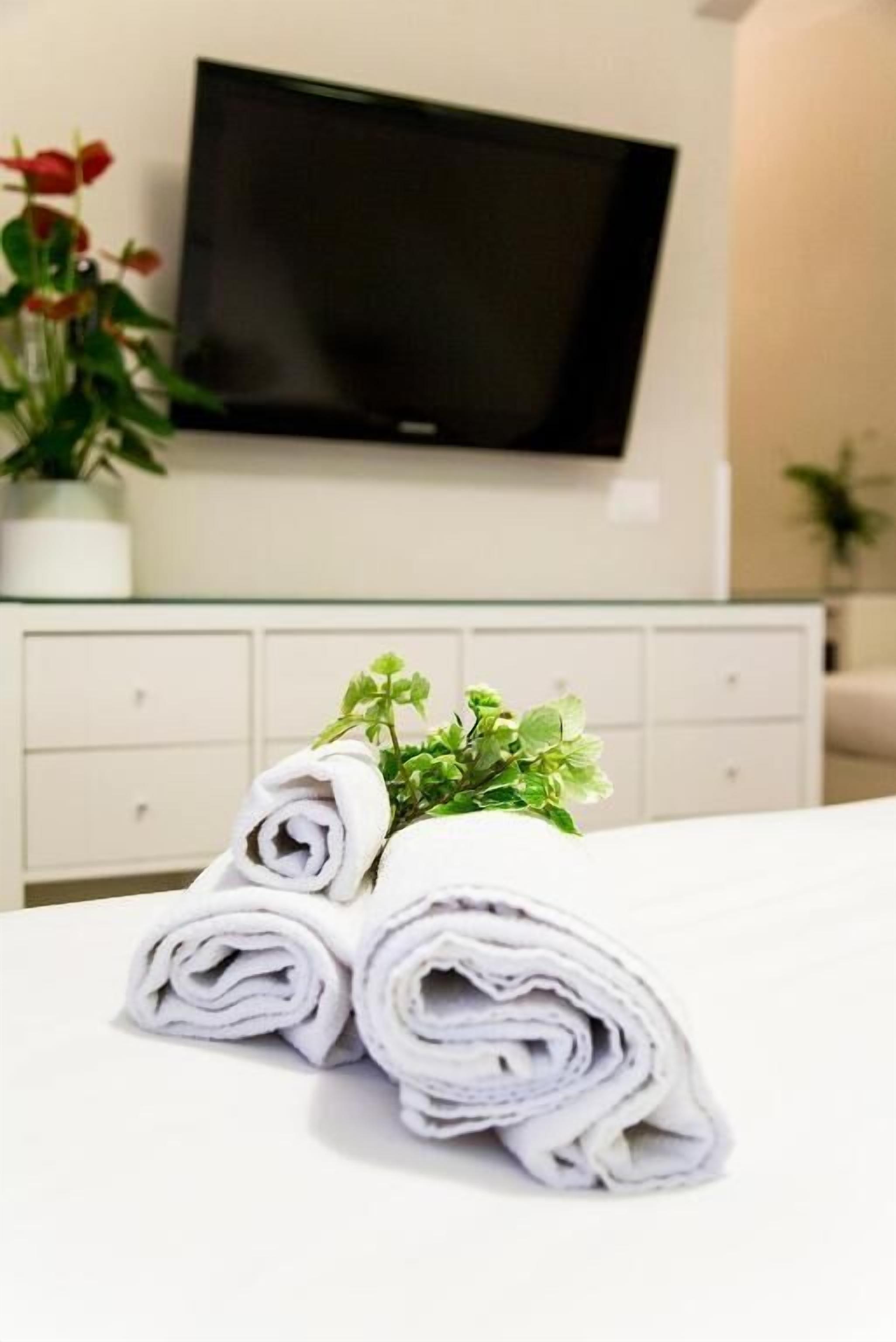 A neatly made bed with rolled white towels and a small green plant on top. A flat-screen TV is mounted on the wall above a white dresser.
