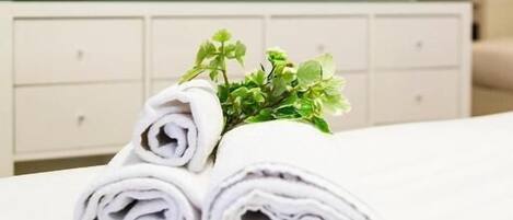 A neatly made bed with rolled white towels and a small green plant on top. A flat-screen TV is mounted on the wall above a white dresser.