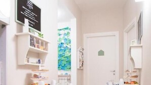 A narrow hallway with white walls and a tiled floor, leading to a door with a small green house symbol. There are two white chairs and a table with various items on it. - B&B Casetta Roma  (Rome)