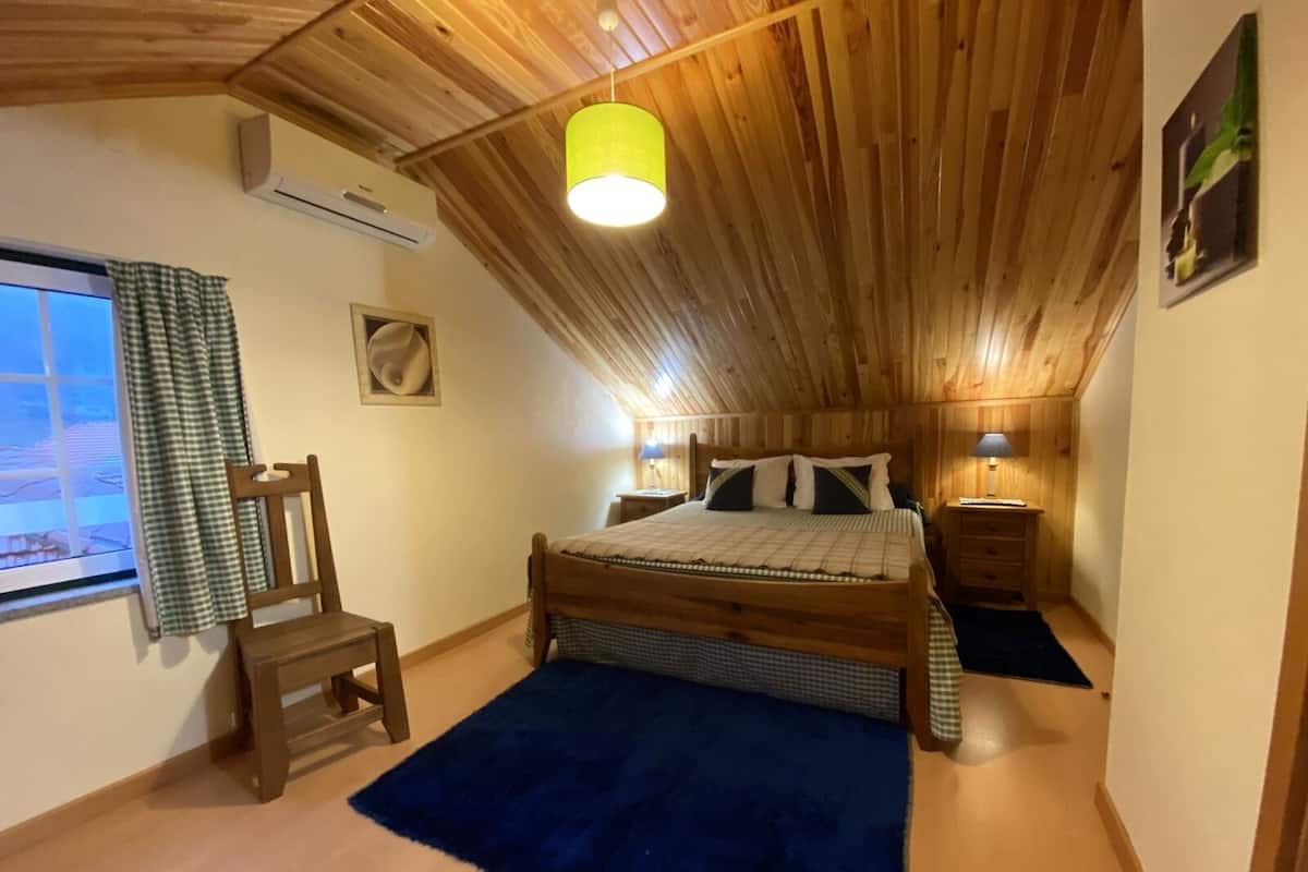 A bedroom with a sloped ceiling, a bed, a chair, a bedside table, and a window with curtains.