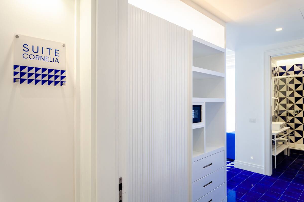 Une chambre d’hôtel avec une pancarte indiquant « SUITE CORNELIA », une armoire blanche, un sol carrelé bleu et un mur orné d’un motif géométrique.