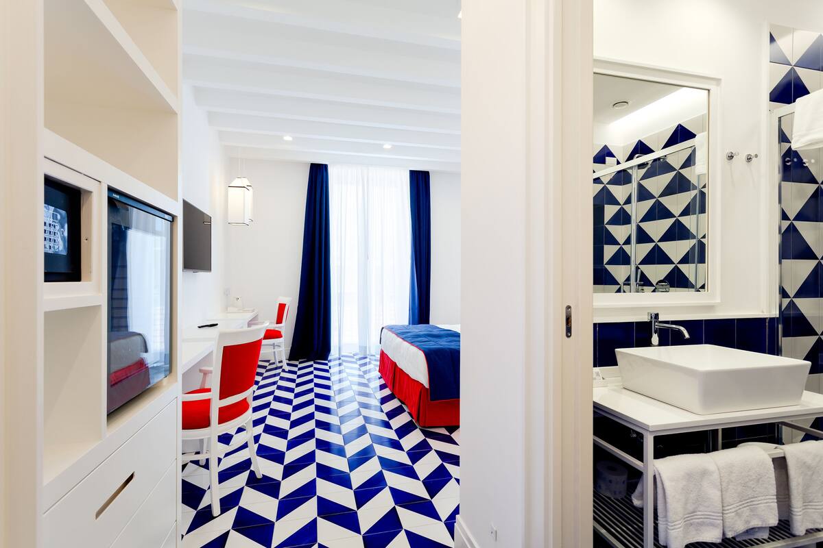 Une chambre d’hôtel moderne avec un sol à motifs géométriques, un lit avec une literie rouge et blanche, un lavabo blanc et une douche aux carreaux bleus et blancs.