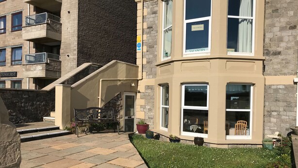 Un edificio de varias plantas con fachada de piedra, una escalera que conduce a la entrada y un pequeño jardín con plantas en macetas.