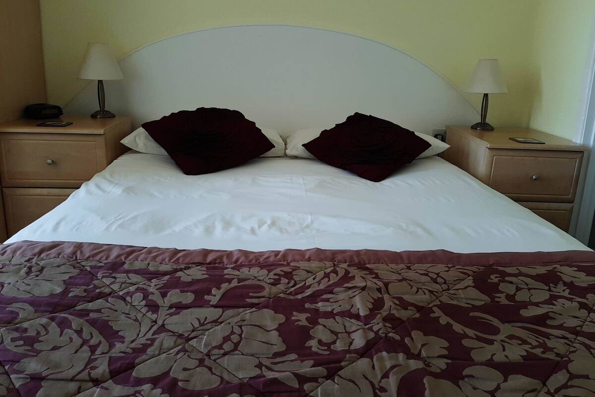 A neatly made bed with a white headboard, two dark red pillows, and a floral-patterned bedspread. There are two bedside tables with lamps and a small clock on one.