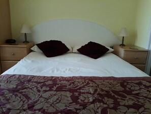 A neatly made bed with a white headboard, two dark red pillows, and a floral-patterned bedspread. There are two bedside tables with lamps and a small clock on one. - The Beaches Family B&B (Weston-super-Mare)