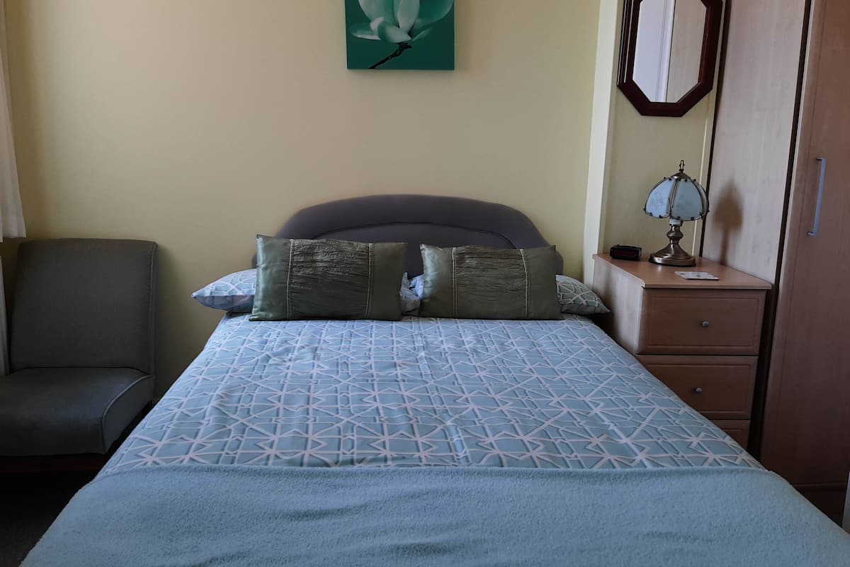 A bedroom with a bed, a nightstand, a lamp, a chair, and a wall-mounted mirror.