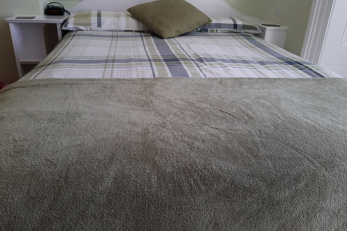 A bed with a plaid bedspread and a green pillow, a bedside table with a lamp, and a power outlet.