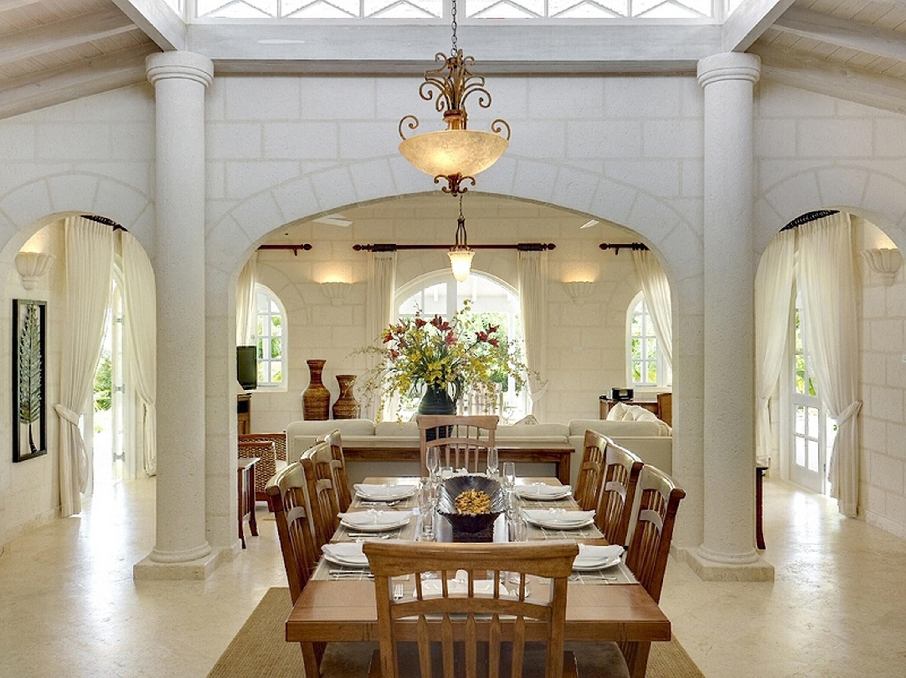 Comedor con techo alto, columnas blancas y una araña. Se observa una mesa de comedor con sillas, y un jarrón con flores sobre la mesa.