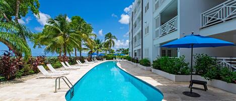 A swimming pool with lounge chairs and umbrellas, surrounded by palm trees and a building with balconies.