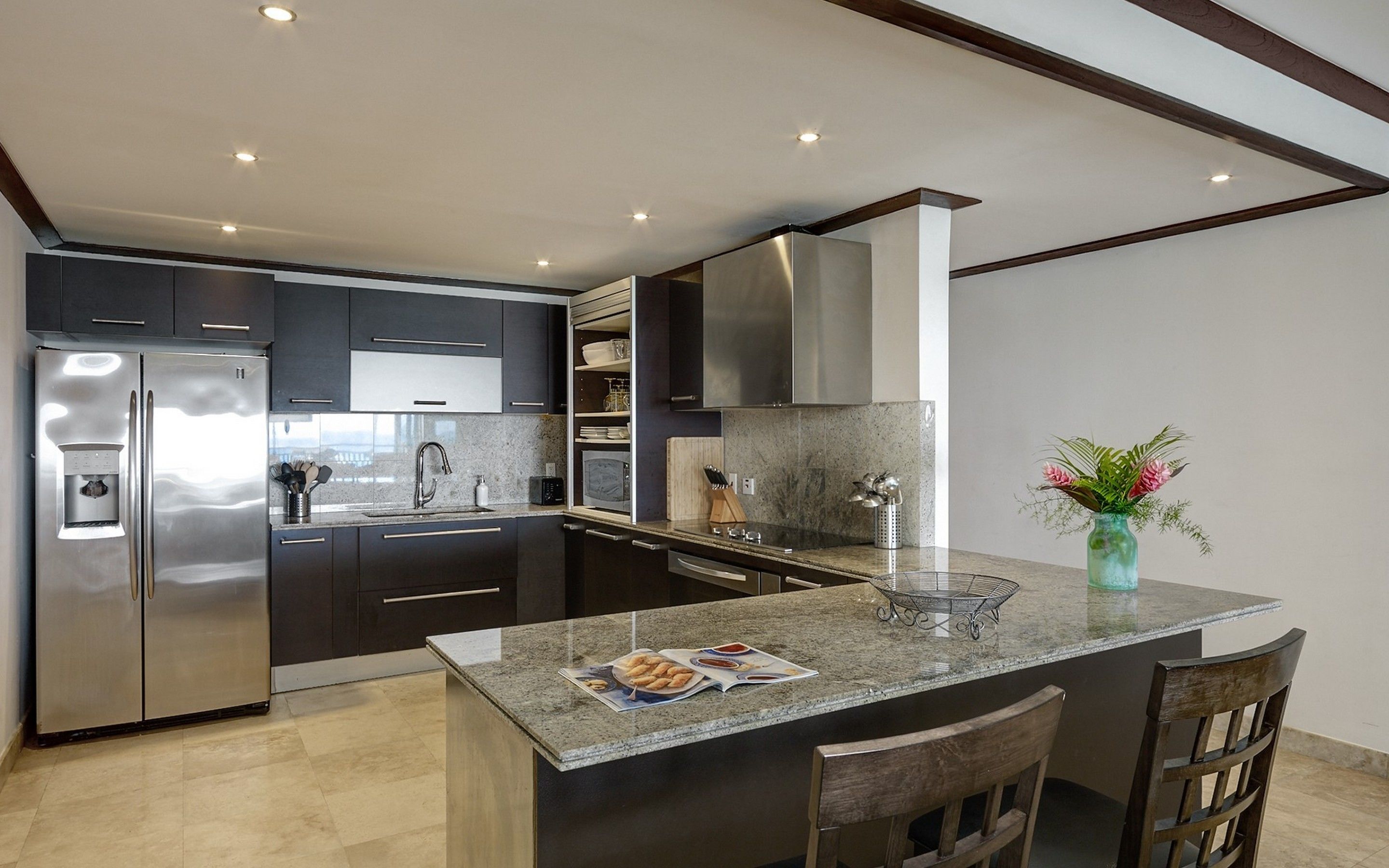 A modern kitchen with a stainless steel refrigerator, dark cabinetry, a granite countertop, and a small island with a vase of flowers.