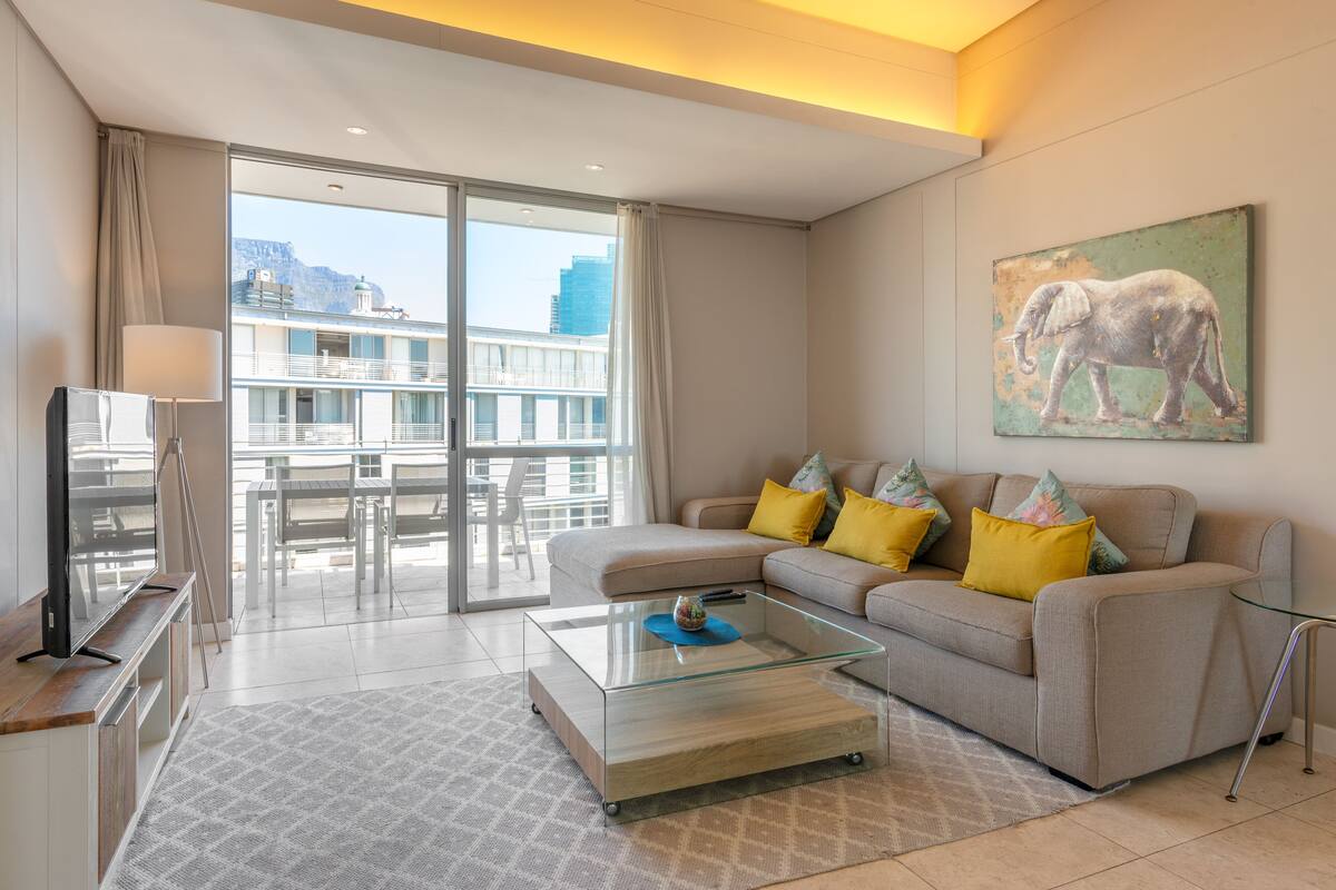 A modern living room with a glass coffee table, a sofa with colorful cushions, a flat-screen TV, and a painting of an elephant on the wall.