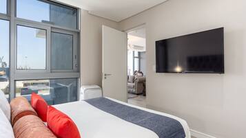 Quarto de hotel moderno com uma cama grande, televisão de tela plana e vista para o exterior por meio de amplas janelas.
