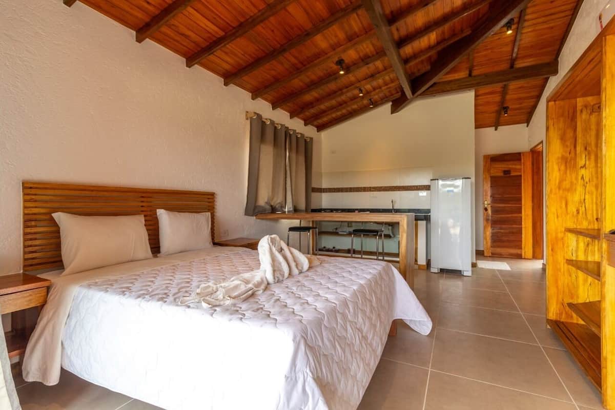 Un dormitorio con cabecera de madera, cama con ropa de cama blanca, un banco de madera y una zona de cocina con refrigerador y sillas.