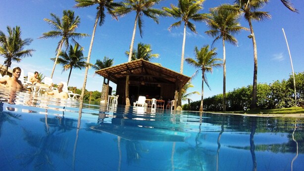A clear blue pool surrounded by palm trees, with a wooden structure and lounge chairs. - Trancoso Residence (Porto Seguro)