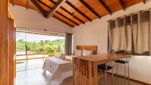 A bedroom with a bed, a wooden table, and two chairs. There is a large window with curtains, and a balcony with a view of greenery. - Trancoso Residence (Porto Seguro)