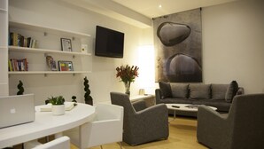 A modern living room with a white round table, grey armchairs, a grey sofa, a flat-screen TV, and a bookshelf with books and decorative items. - Hostal Goya (Barcelona)