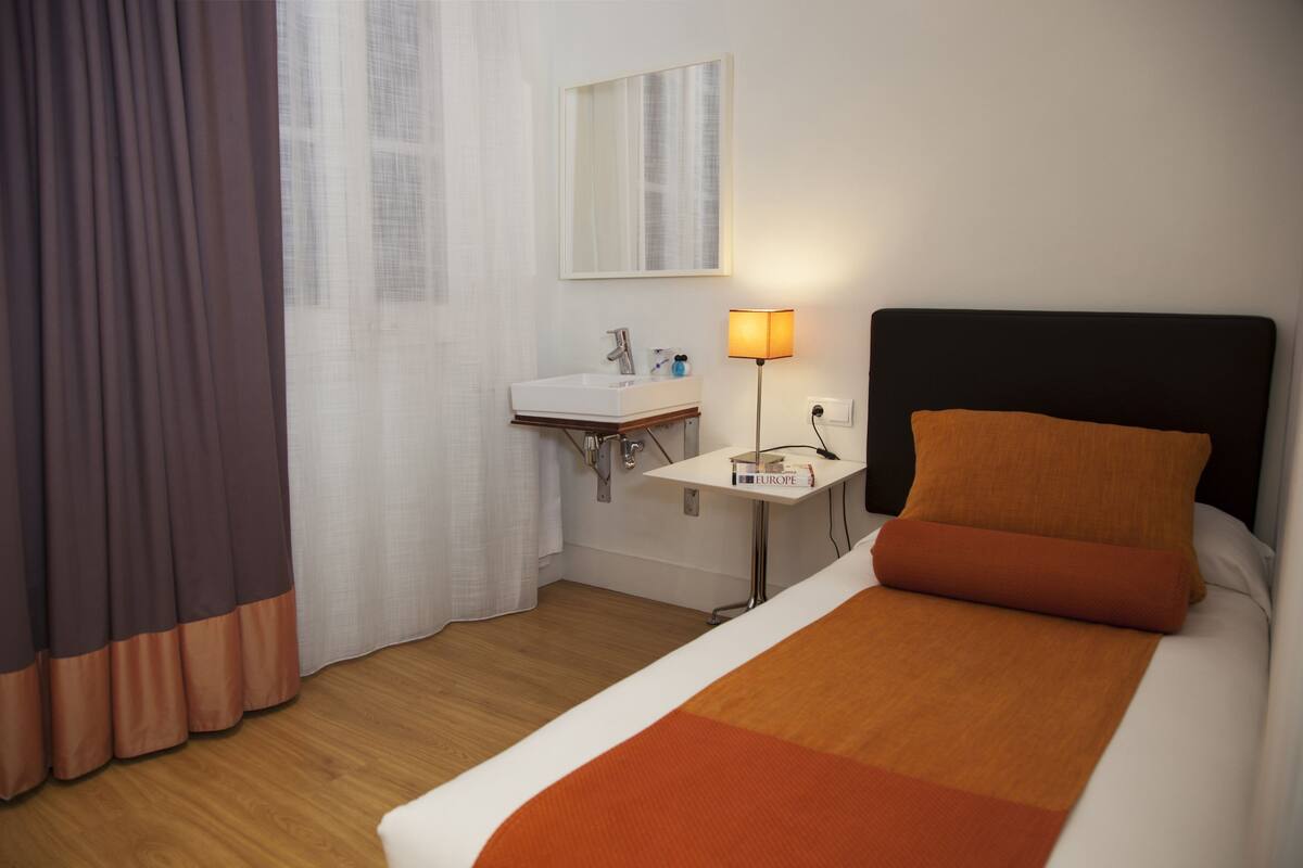 A hotel room with a bed, a bedside table with a lamp, a small bathroom with a sink, and a window with curtains.