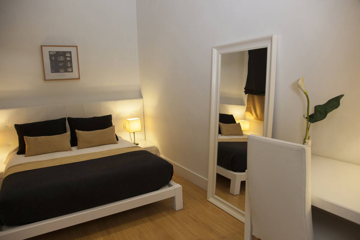 A modern bedroom with a bed, bedside lamps, a mirror, and a plant.