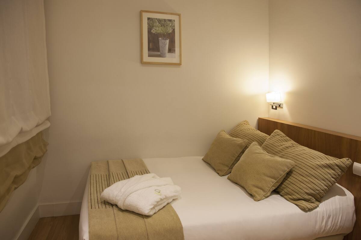 A neatly made bed with a beige blanket and pillows, a white towel, and a framed picture on the wall.