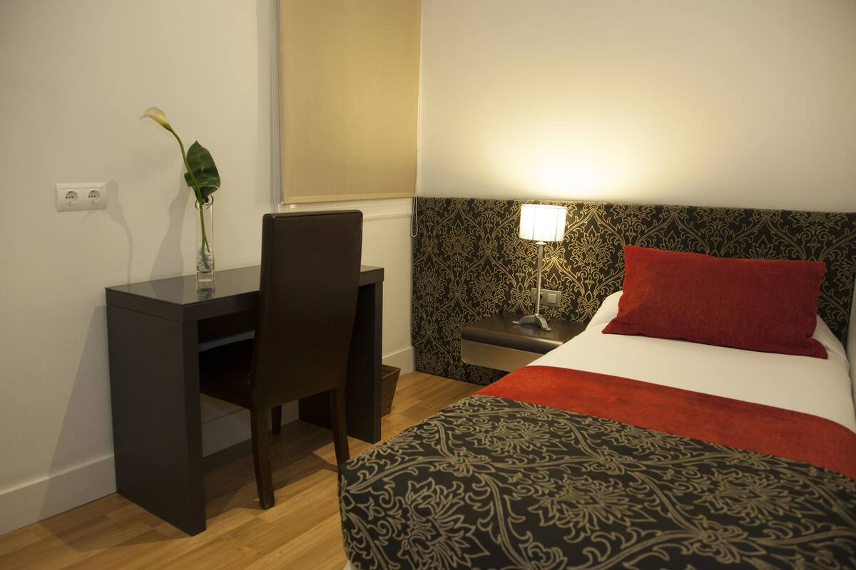 A hotel room with a bed, a desk, a chair, a nightstand, a lamp, and a plant.