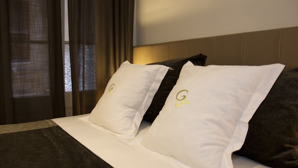 A hotel room with a bed featuring white and dark gray pillows with a logo, a headboard, and a window with curtains. - Hostal Goya (Barcelona)