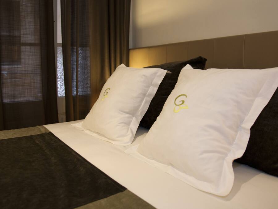 Habitación de hotel con una cama, almohadas blancas y gris oscuro con un logotipo, cabecera y ventana con cortinas.