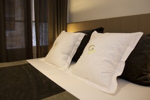 A hotel room with a bed featuring white and dark gray pillows with a logo, a headboard, and a window with curtains. - Hostal Goya (Barcelona)