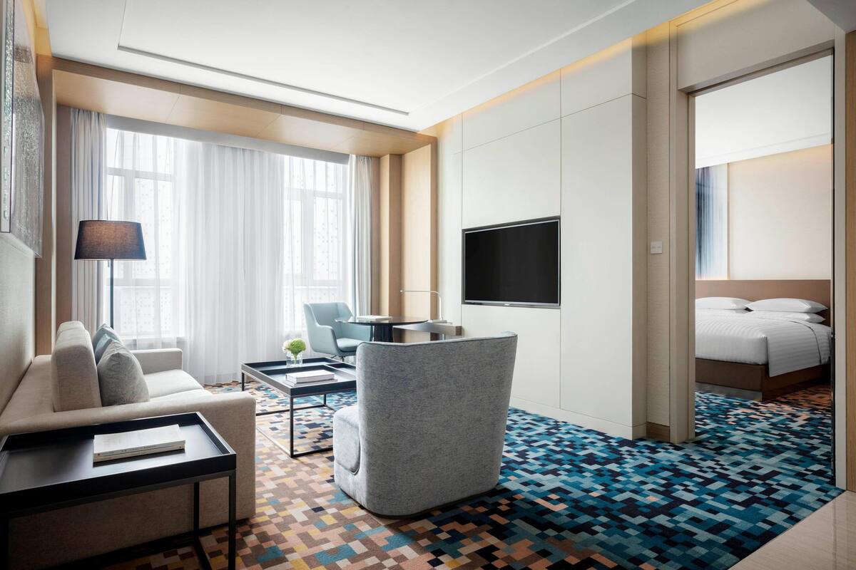 A modern hotel room with a sofa, armchair, flat-screen TV, and a bed.