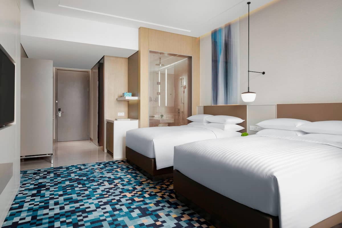 A hotel room with two beds, a patterned rug, a wooden headboard, a bedside table, and a wall-mounted light fixture.