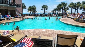 A swimming pool with lounge chairs and umbrellas, surrounded by palm trees and a building with a balcony. - Long Bay 216 (Myrtle Beach)