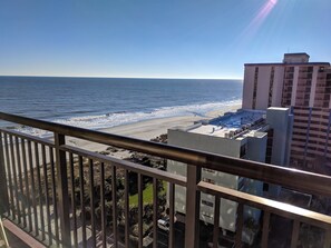 A balcony with a view of the beach and ocean, a parking area, and a building with multiple floors. - Caravelle 1401 (Myrtle Beach)