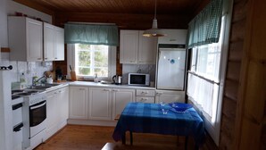 Une cuisine compacte avec des meubles blancs, une cuisinière, un four à micro-ondes et un réfrigérateur. On y trouve une fenêtre avec des rideaux à carreaux, une table recouverte d’une nappe bleue et un banc en bois.